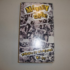May include: A VHS video cassette with a collage of black and white photos of the band Motley Crue. The title "Motley Crue" is in yellow letters on a black background. The text "Decade of Decadence '81-'91" is in yellow letters on a black background.
