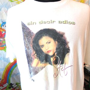 Könnte beinhalten: Weißes T-Shirt mit einem Porträt einer Frau mit dunklem Haar und rotem Lippenstift. Das Shirt hat den Text "Sin decir adios" über dem Bild und eine Unterschrift darunter. Das Shirt befindet sich auf einer Schaufensterpuppe.