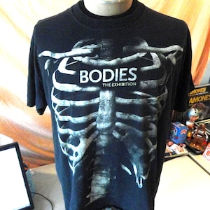 May include: Black t-shirt featuring a white skeletal rib cage design. The text "BODIES THE EXHIBITION" is printed across the top of the rib cage. The shirt is displayed on a mannequin.