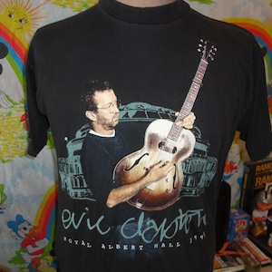 Op de afbeelding: Zwart T-shirt met een afbeelding van Eric Clapton die gitaar speelt. De tekst "Royal Albert Hall 1991" staat onder de afbeelding.
