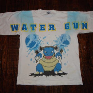 Puede incluir: Camiseta blanca con un gráfico azul y amarillo de un personaje Pokemon, Blastoise, disparando agua de dos cañones. El texto "WATER GUN" está impreso en letras azules y amarillas en la parte superior de la camiseta.