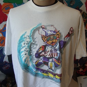 May include: White t-shirt with a graphic of a cartoon bear wearing a purple hat and goggles snowboarding down a wave. The bear is wearing a red, white, and blue shirt with "U.S. Snowboarding" written on it.