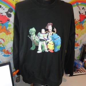 Może przedstawiać: Czarna bluza z długim rękawem z nadrukiem postaci z serii film&oacute;w Toy Story. Postacie to Woody, Buzz Astral, Rex i fioletowy kosmita.