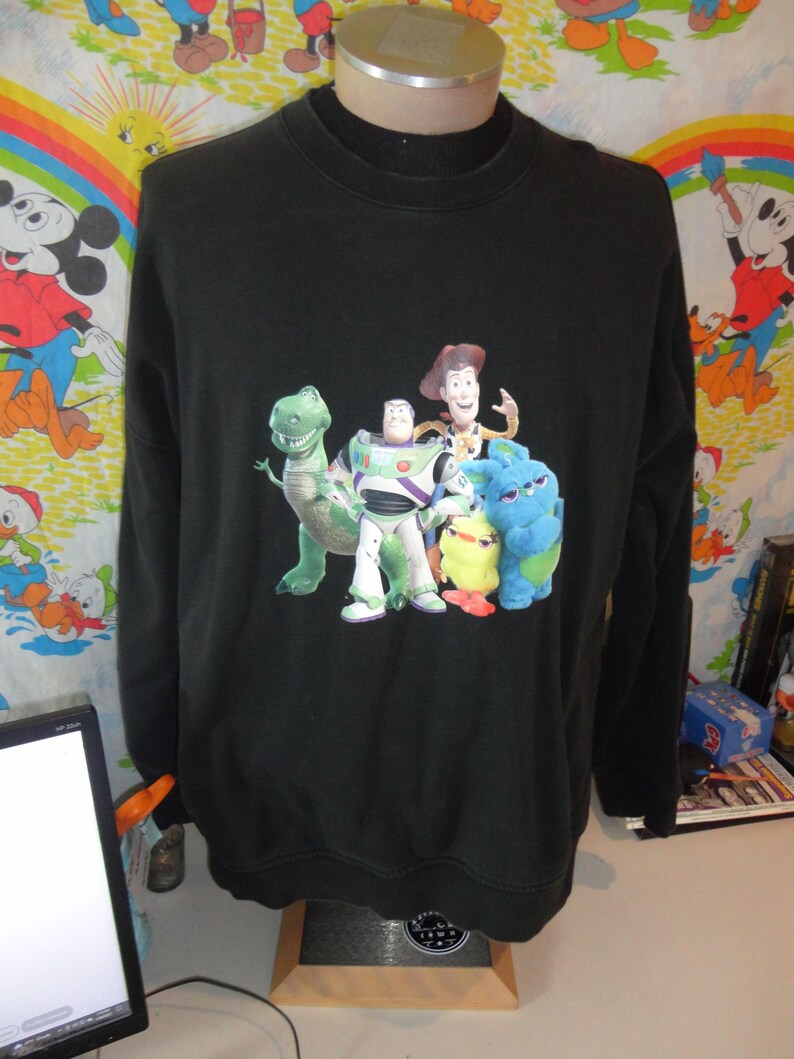 Może przedstawiać: Czarna bluza z długim rękawem z nadrukiem postaci z serii film&oacute;w Toy Story. Postacie to Woody, Buzz Astral, Rex i fioletowy kosmita.