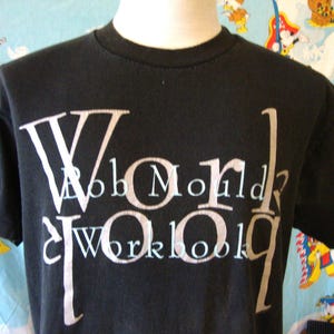 May include: A black short-sleeve t-shirt featuring large, stylized white lettering that reads "WORK" vertically and horizontally, with the text "BOB MOULD" and "WORKBOOK" printed in a smaller, lighter font across the center of the chest.