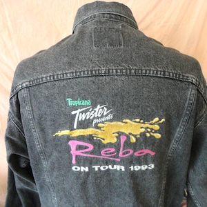 Op de afbeelding: Een verbleekte zwarte spijkerjas met de tekst "Tropicana Twister presents Reba ON TOUR 1993" geborduurd op de rug in groen, wit, geel en roze. De jas heeft een klassieke kraag en een knoopsluiting.