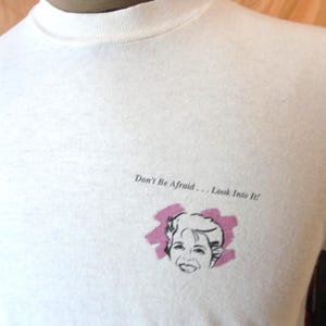 May include: White t-shirt featuring a black and white illustration of a woman's face within a pink oval. The text "Don't Be Afraid... Look Into It!" is printed above the illustration. The shirt has a crew neck.