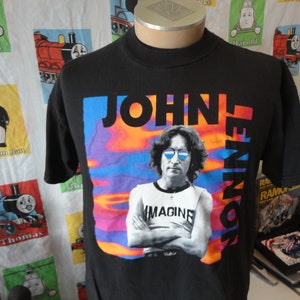 May include: Black t-shirt with a colorful image of John Lennon wearing sunglasses and a white t-shirt that says "Imagine". The text "John Lennon" is printed vertically on the right side of the image.