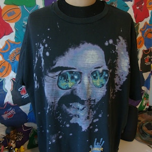 Puede incluir: Camiseta negra con una imagen descolorida y colorida de un hombre con gafas de sol. La imagen est&aacute; rodeada de pintura blanca salpicada. El texto "Remarkable" est&aacute; impreso en la parte inferior de la camiseta.