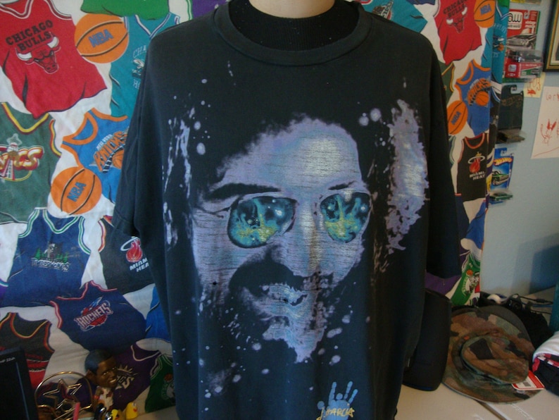 Puede incluir: Camiseta negra con una imagen descolorida y colorida de un hombre con gafas de sol. La imagen est&aacute; rodeada de pintura blanca salpicada. El texto "Remarkable" est&aacute; impreso en la parte inferior de la camiseta.