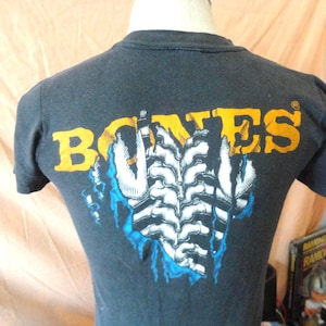 May include: Dark gray t-shirt with the word "BONES" in yellow and orange block letters. Below the word "BONES" is a white and black graphic design with blue accents. The t-shirt is displayed on a mannequin.