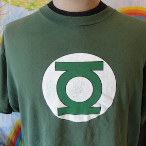 May include: Green t-shirt featuring the Green Lantern logo, a white circle with a green emblem. The shirt has a crew neck and short sleeves. The shirt is a dark green color.