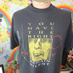 May include: Black t-shirt with the text "YOU HAVE THE RIGHT" in yellow block letters. Below the text is a yellow image of a face. The words "NOT TO REMAIN SILENT?" are printed below the image.