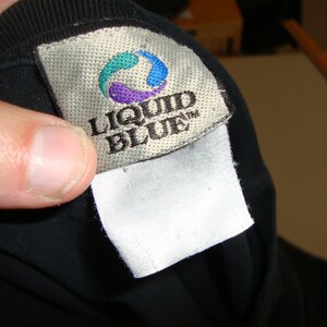 Puede incluir: Una camiseta negra con una etiqueta blanca que dice "Liquid Blue" en letras negras. La etiqueta tiene un logotipo azul, verde y morado.