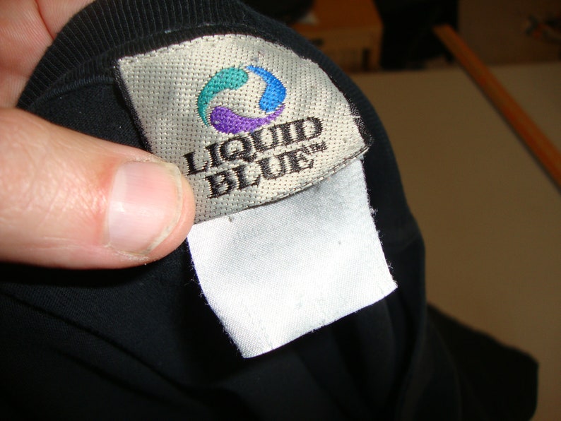 Puede incluir: Una camiseta negra con una etiqueta blanca que dice "Liquid Blue" en letras negras. La etiqueta tiene un logotipo azul, verde y morado.