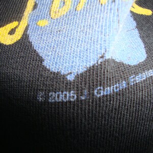 Puede incluir: Primer plano de una camiseta negra con un dise&ntilde;o gr&aacute;fico azul y amarillo. El texto "J. Garcia Escaio" est&aacute; impreso en la parte inferior de la camiseta.