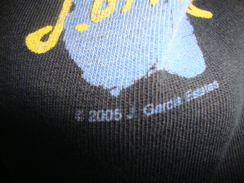 Puede incluir: Primer plano de una camiseta negra con un dise&ntilde;o gr&aacute;fico azul y amarillo. El texto "J. Garcia Escaio" est&aacute; impreso en la parte inferior de la camiseta.