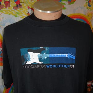 Puede incluir: Camiseta negra con un gráfico de una guitarra eléctrica blanca y azul sobre un fondo azul y verde. El texto "ERIC CLAPTON WORLD TOUR/01" está impreso debajo de la imagen de la guitarra. La camiseta se muestra en un maniquí.