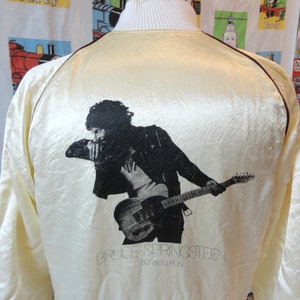 May include: A cream-colored satin bomber jacket with a black and white image of Bruce Springsteen playing a guitar. The text "Bruce Springsteen Born to Run" is printed below the image.