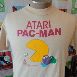 Puede incluir: Una camiseta beige con un gráfico de Pac-Man amarillo y el texto "ATARI PAC-MAN" en letras rosa y rojo. La camiseta también incluye un pequeño logotipo de Atari.