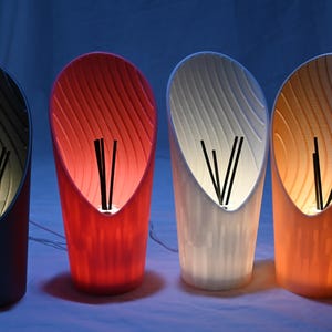 May include: Four modern lamps in a row, each with a different colour: dark grey, red, white, and orange. The lamps have a curved, open design with a textured interior and black accents.
