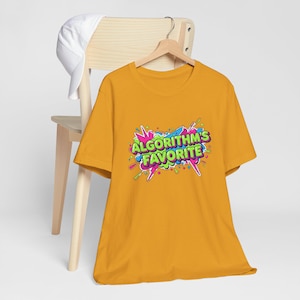 Könnte beinhalten: Senfgelbes T-Shirt mit dem Text "ALGORITHM'S FAVORITE" in einem farbenfrohen Comic-Design. Das Shirt hängt an einem Holzkleiderbügel, mit einer weißen Baseballkappe auf einem Holzstuhl im Hintergrund.