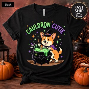 May include: A black t-shirt featuring a graphic of a cartoon corgi wearing a witch hat and cape, stirring a bubbling green cauldron. The text "CAULDRON CUTIE" is printed in an arched design above the dog, surrounded by small colorful stars.