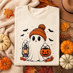 May include: A cream-colored t-shirt featuring a graphic of a white ghost wearing an orange knit beanie. The ghost holds a dark gray lantern in one hand and an orange jack-o'-lantern candy bucket in the other, surrounded by small black bats.