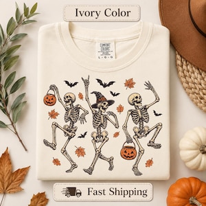May include: An ivory-colored t-shirt featuring a graphic of three dancing skeletons. One skeleton wears a witch hat, while two hold jack-o'-lantern buckets. The design includes black bats and orange autumn leaves. The shirt has a Comfort Colors label.
