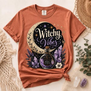 May include: A rust-colored t-shirt featuring a graphic print of a crescent moon, purple crystals, and a glass bottle with a label that reads "MAGIC WITHIN". The text "Witchy Vibes" is printed in a decorative font above the bottle illustration.