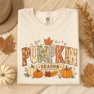 Pode incluir: Uma t-shirt de cor creme com o texto PUMPKIN SEASON em letras grandes preenchidas com vários padrões de outono. Abaixo do texto, encontram-se ilustrações de duas abóboras, folhas de outono e pequenas flores em tons de laranja, castanho e verde.