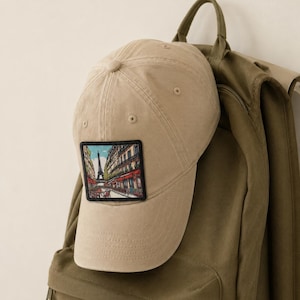 May include: A beige baseball cap featuring a square patch on the front panel. The patch displays a colorful illustration of a Parisian street scene with the Eiffel Tower in the background, framed by buildings and outdoor cafe seating with red awnings.