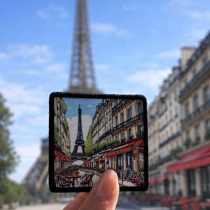 May include: A square patch featuring a street scene in Paris with the Eiffel Tower in the background. The patch has a black border and the word "Paris" is written in cursive. The background shows the Eiffel Tower and a blue sky.