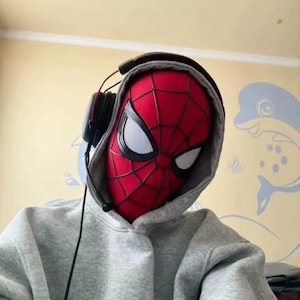 Op de afbeelding: Een rood en zwart Spider-Man-masker met grote witte ooglenzen, gedragen onder een grijze hoodie. Een zwarte over-ear koptelefoon met een dunne microfoonarm is over de capuchon geplaatst, met een zwarte draad die naar beneden loopt.
