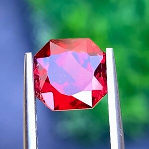 May include: A faceted, octagonal-cut, deep red gemstone held by silver tweezers. The gem has a brilliant, reflective surface, showcasing internal color variations. The background is a blurred mix of green and blue.