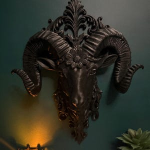 May include: A black ram's head wall decoration with intricate detailing, including curled horns and floral embellishments. The ram's head is mounted on a dark green wall, with a candle and a potted succulent in the foreground.