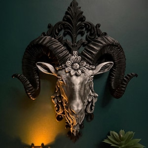 May include: A silver and black ram head wall sculpture with large curved horns and a floral crown. The ram head is mounted on a dark green wall. A decorative candle holder and a potted succulent are in the foreground.