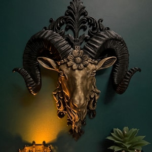 May include: A decorative ram's head wall sculpture with black horns and a gold-toned face, featuring a floral crown. The sculpture is mounted on a dark green wall. A candle holder and a potted succulent are in the foreground.