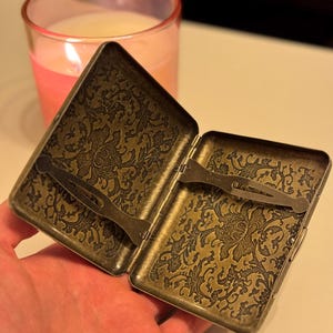 Könnte beinhalten: Eine offene Zigarettenetui im antiken Stil mit einem verzierten Blumenmuster in Bronze- und Goldtönen. Das Etui hat einen Verschluss und einen Metallclip im Inneren. Eine brennende rosa Kerze ist im Hintergrund.