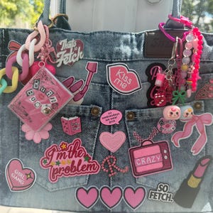 Può includere: Una borsa tote in denim blu decorata con numerosi toppe e ciondoli a tema rosa. Il testo visibile include "That's So Fetch", "Kiss me", "on wednesdays we wear pink", "I'm the problem", "CRAZY", "GIRL GANG", "BURN BOOK" e "SO FETCH".