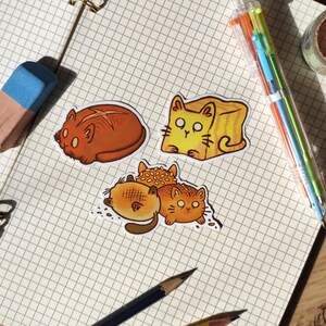 Könnte beinhalten: Eine Sammlung von Aufklebern zum Thema Katze auf kariertem Papier. Die Aufkleber zeigen Cartoon-Katzen, die wie Brot gestaltet sind, darunter ein Laib, eine Toastscheibe und eine Gruppe von katzenförmigem Gebäck. Ein Bleistift, ein Radiergummi und Stifte sind ebenfalls zu sehen.