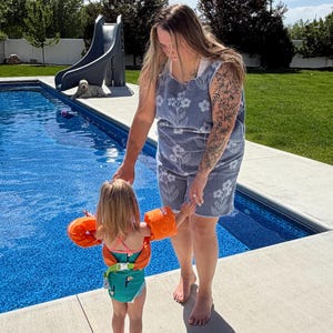 May include: A blue terry cloth tank top and shorts set featuring a white floral pattern. The set is worn with a green swimsuit featuring a toucan print and bright orange inflatable arm floaties with a white buckle strap at the waist.