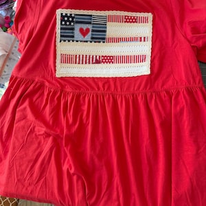 May include: A bright red t-shirt featuring a rectangular patchwork applique on the chest. The applique resembles an American flag made from various red, white, and blue fabric scraps, accented with a red heart and white lace trim borders.