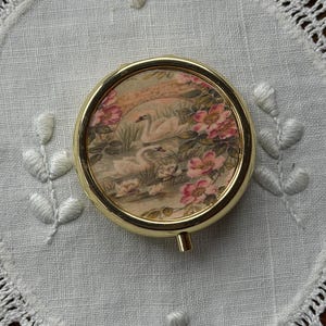 May include: A round gold-tone metal compact mirror featuring a decorative insert on the lid. The insert displays a printed illustration of two white swans swimming in a pond surrounded by pink flowers and green foliage under a stone bridge.