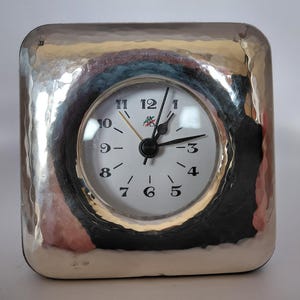 Puede incluir: Un reloj analógico de forma cuadrada con un marco de metal plateado pulido y martillado. La esfera es blanca con números arábigos negros del 1 al 12, manecillas negras para horas y minutos, un segundero dorado fino y un pequeño logotipo.