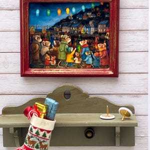Könnte beinhalten: Ein gerahmtes Kunstwerk zeigt anthropomorphe Mäuse in einem Dorf bei Nacht, beleuchtet von Laternen. Darunter hängt ein Weihnachtsstrumpf voller Geschenke in der Nähe eines kleinen Regals mit dekorativen Gegenständen.