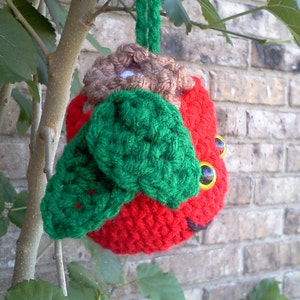 Op de afbeelding: Een rood gehaakt lieveheersbeestje ornament met groene vleugels en zwarte ogen. Het lieveheersbeestje hangt aan een bruin touwtje.