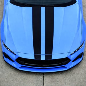 May include: A bright blue car hood featuring two bold, vertical matte black racing stripes running from the windshield to the front bumper, complemented by a black front grille and sharp, angular LED headlight accents on both sides.