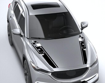 May include: A silver SUV featuring two black and white geometric hood decals. The decals consist of parallel stripes and square pixel-like patterns that extend from the base of the windshield toward the front grille of the vehicle.