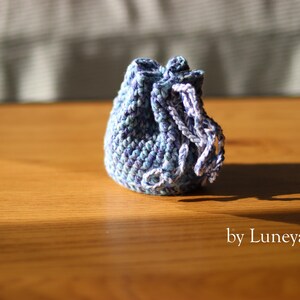 May include: A small, handmade crochet pouch in shades of blue and white, with a drawstring closure. The pouch is sitting on a wooden surface. The text "by Luneya.shop" is visible.